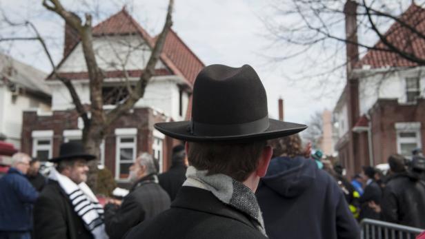 Eine Menschenmenge, darunter Männer mit traditionellen jüdischen Hüten, steht auf einer Straße.