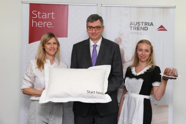 Eine Frau mit Kissen, ein Mann im Anzug und ein Zimmermädchen mit Kuchen vor einem Austria Trend Hotels Banner.