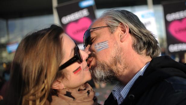 Ein Paar mit aufgemalten Flaggen küsst sich vor Plakaten mit der Aufschrift „Griechenland“.