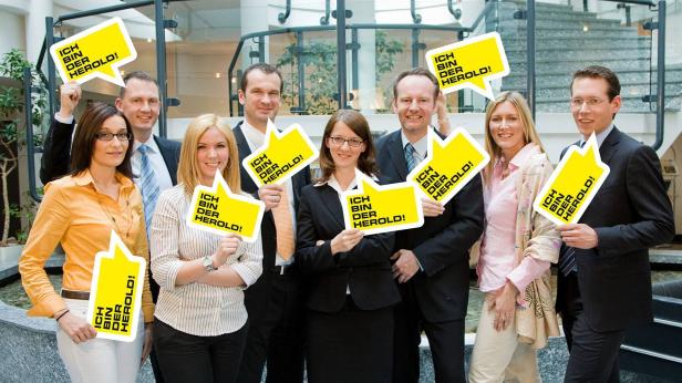 Eine Gruppe von Büroangestellten hält gelbe Schilder mit der Aufschrift „Ich bin der Herold!".