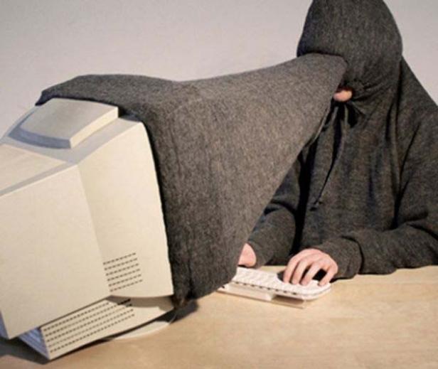 Eine Person mit Kapuze ist durch einen grauen Stoff mit einem alten Computermonitor verbunden und tippt auf einer Tastatur.