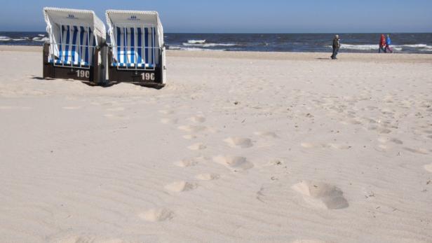 Zwei Strandkörbe stehen am Strand, im Hintergrund spazieren Menschen am Ufer entlang.