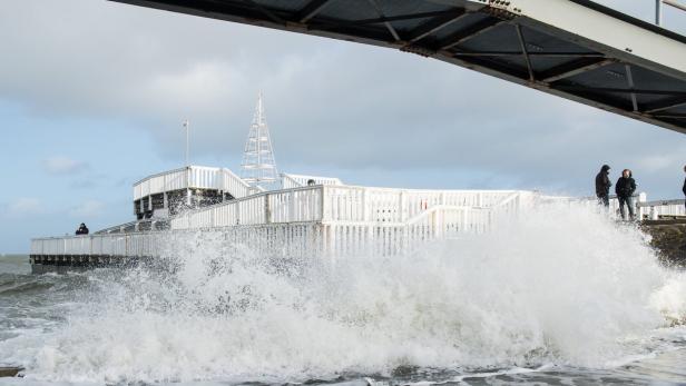 Eine große Welle bricht an einem Pier mit Menschen und einem dekorativen Turm.
