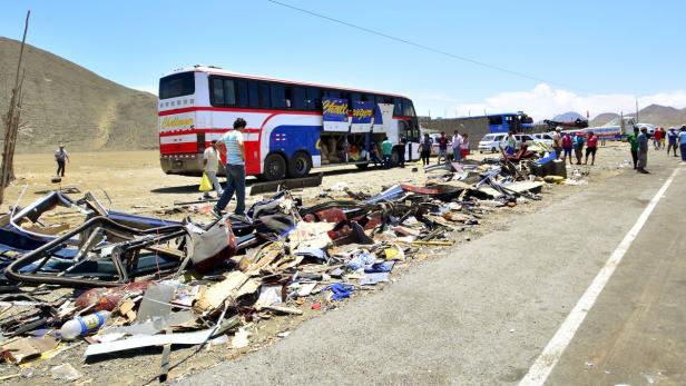 Ein zerstörter Bus steht neben einer Straße, umgeben von Trümmern und Menschen.