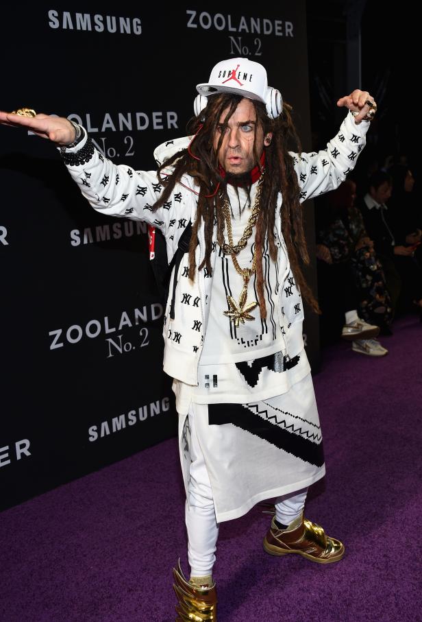 Ein Mann mit Dreadlocks, Mütze und auffälligen goldenen Schuhen posiert auf einem lila Teppich.