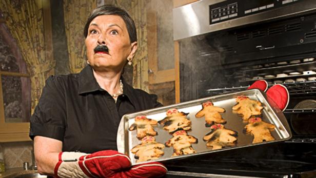 Eine Frau mit aufgemaltem Schnurrbart hält ein Backblech mit verbrannten Lebkuchenmännern vor einem Ofen.