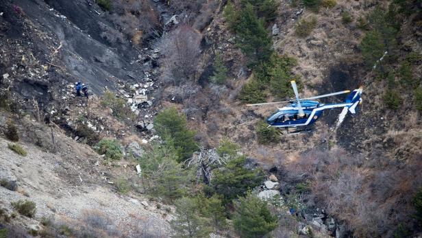 Ein Hubschrauber fliegt über eine Bergflanke mit Trümmerteilen und zwei Personen.