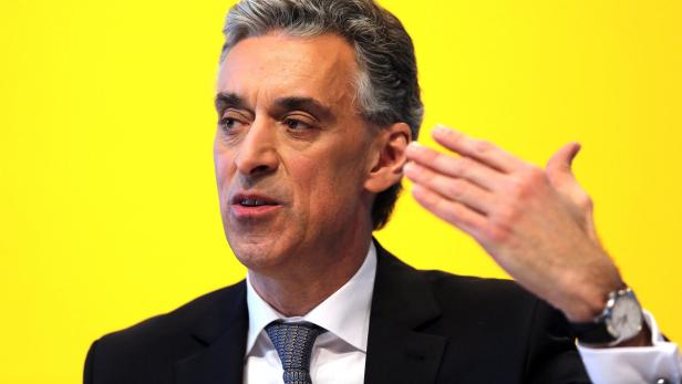 Ein Mann in Anzug gestikuliert vor einem gelben Hintergrund.