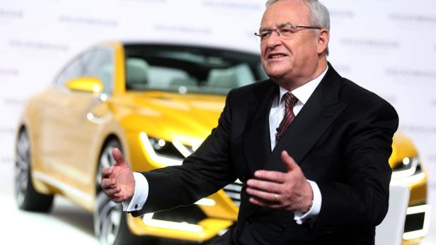 Ein Mann in einem Anzug gestikuliert vor einem gelben Auto.