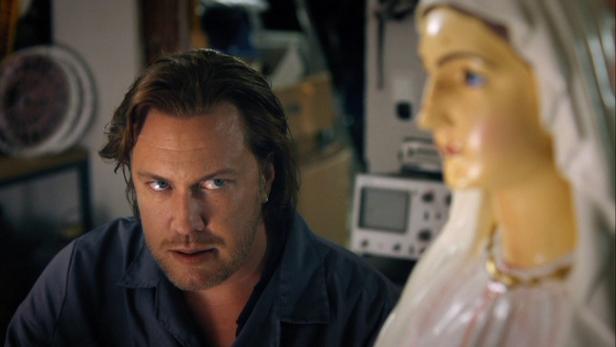 Ein Mann mit Bart und blauen Augen sitzt neben einer Marienstatue.