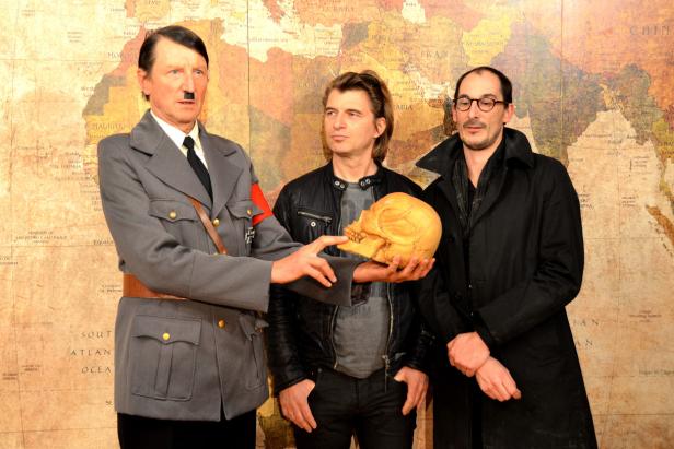 Drei Männer posieren vor einer Weltkarte, einer hält einen Schädel, einer ist als Hitler verkleidet.