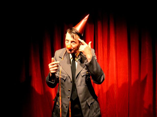 Ein Mann mit Clownsnase und Partyhut in Uniform vor einem roten Vorhang.