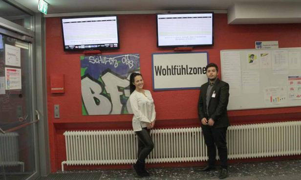 Eine Frau und ein Mann stehen vor einer Wand mit Graffiti und Bildschirmen.