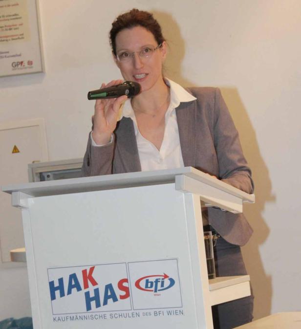 Eine Frau mit Brille hält ein Mikrofon an einem Rednerpult mit dem Logo von HAK/HAS und BFI Wien.