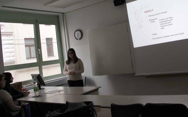 Eine junge Frau präsentiert ihr Projekt vor einer kleinen Gruppe in einem Klassenzimmer.