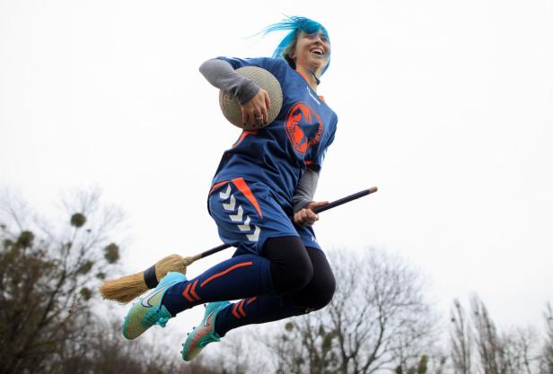 Eine Frau mit blauen Haaren springt auf einem Besen und hält einen Ball.