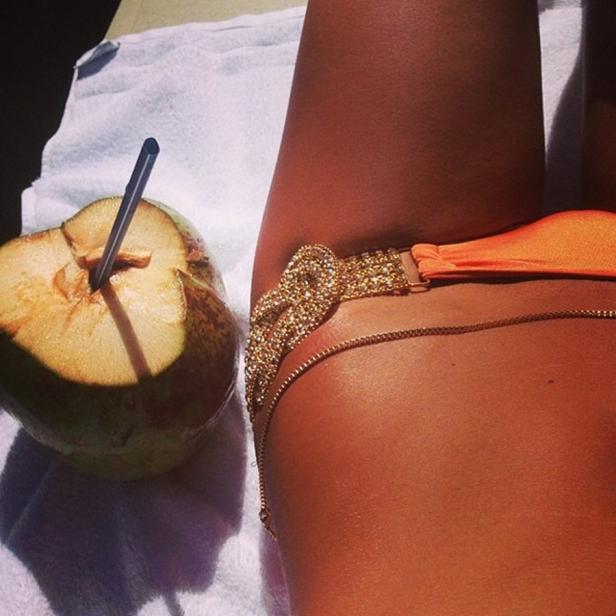 Eine Frau liegt mit einem Getränk in einer Kokosnuss und einem goldenen Beinschmuck am Strand.