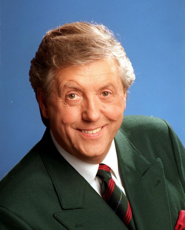 Ein lächelnder Mann im grünen Anzug mit Krawatte vor blauem Hintergrund.