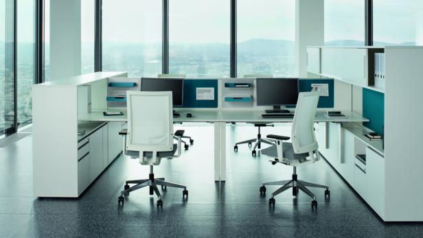 Ein modernes Büro mit weißen Schreibtischen, Bürostühlen und Computern vor einem großen Fenster.