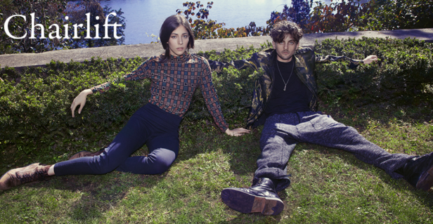 Die Band Chairlift sitzt auf einer Wiese vor einem See.