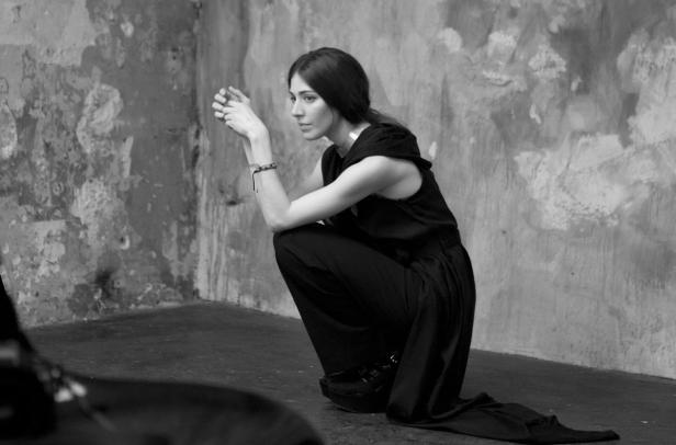 Eine Frau in dunkler Kleidung kauert vor einer strukturierten Wand.