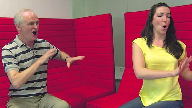 Ein Mann und eine Frau sitzen auf roten Stühlen und gestikulieren mit geöffnetem Mund.