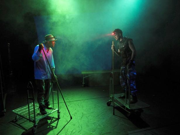 Zwei Schauspieler stehen auf Rollwagen unter grünem Licht und Nebel auf einer Bühne.