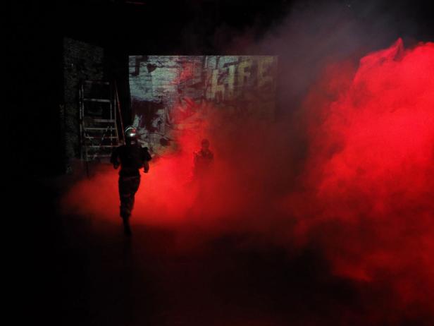 Eine Person rennt durch roten Rauch vor einer Wand mit Graffiti.