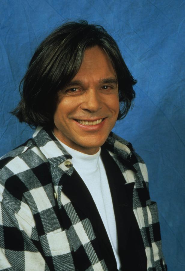 Ein lächelnder Mann mit langem Haar und kariertem Hemd vor blauem Hintergrund.