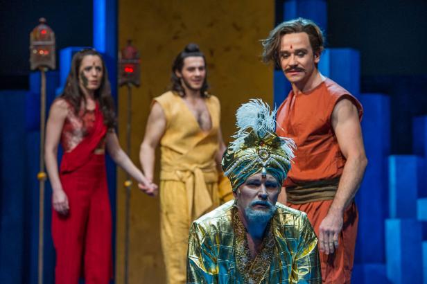 Eine Theaterszene mit Darstellern in farbenfrohen Kostümen vor einem Bühnenbild.