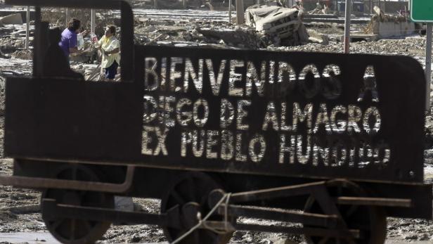 Ein Schild mit der Aufschrift „Bienvenidos a Diego de Almagro, ex pueblo hundido“ vor einer zerstörten Landschaft.