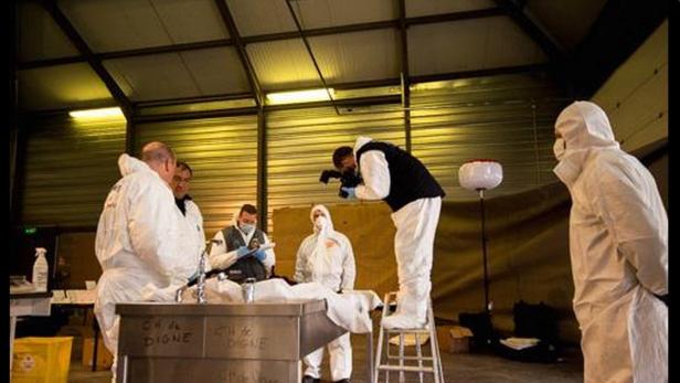 Mehrere Personen in Schutzanzügen untersuchen einen Tatort in einer Lagerhalle.
