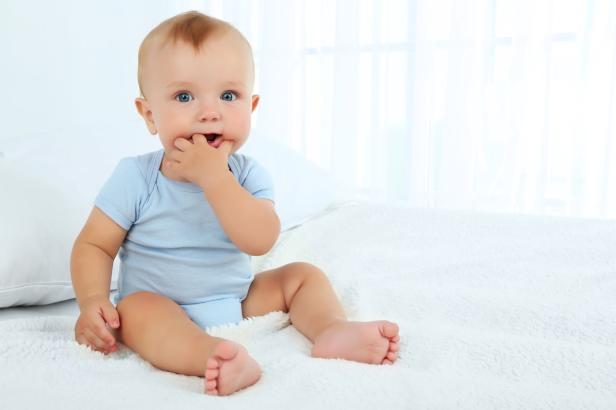 Baby, Bub Ein blau gekleideter Säugling sitzt auf einer weißen Decke und lutscht an seinen Fingern.