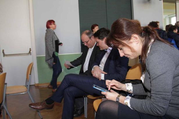 Eine Frau tippt auf einem Tablet, während andere Personen im Hintergrund sitzen oder stehen.