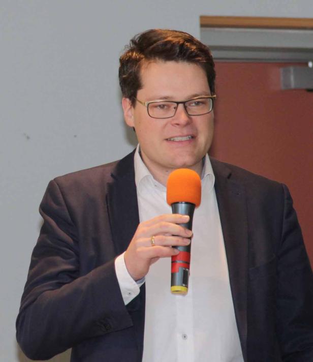 Ein Mann mit Brille und Anzug spricht in ein orangefarbenes Mikrofon.