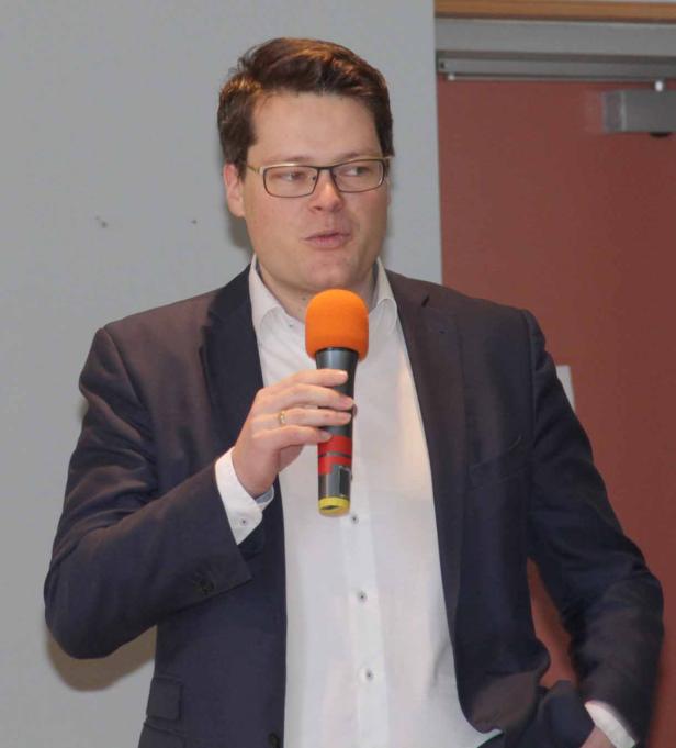 Ein Mann mit Brille und Anzug spricht in ein orangefarbenes Mikrofon.