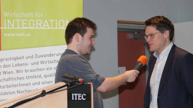 Ein Mann interviewt einen anderen Mann mit einem orangefarbenen Mikrofon vor einem Banner für „Wirtschaft für Integration“.