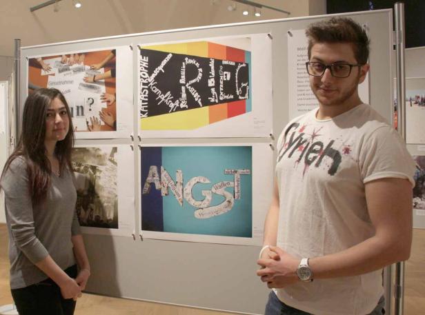 Zwei Personen stehen vor einer Ausstellung mit Plakaten, die Wörter wie „Angst“ und „Katastrophe“ zeigen.