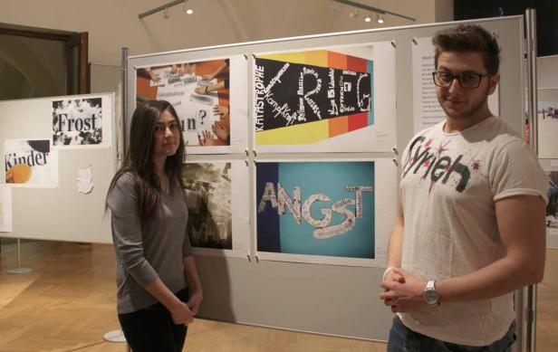 Zwei Personen stehen vor einer Ausstellung mit Plakaten, die Wörter wie „Angst“ und „Katastrophe“ zeigen.