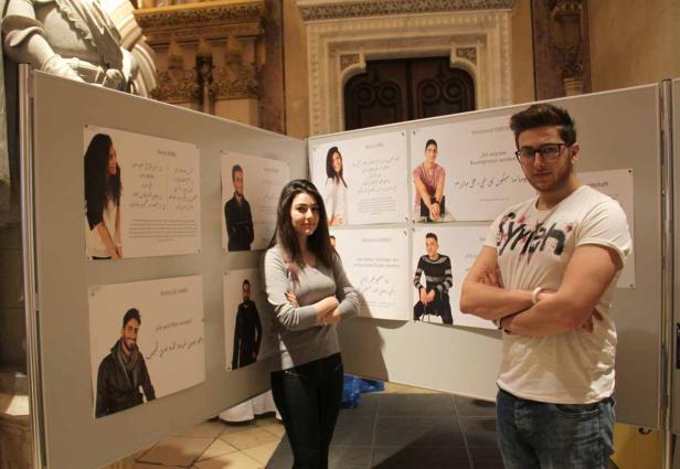 Eine junge Frau und ein junger Mann stehen vor einer Ausstellung mit Porträts und Texten.
