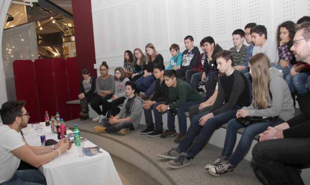 Eine Gruppe Jugendlicher sitzt auf einer Treppe und hört einem Mann an einem Tisch zu.