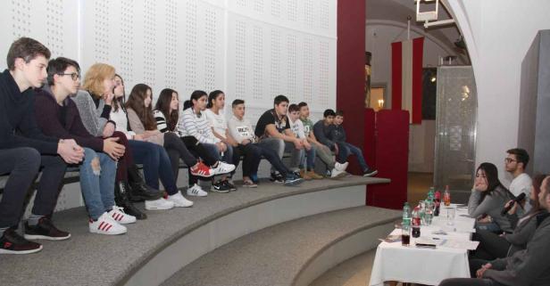 Eine Gruppe Jugendlicher sitzt auf einer Treppe und hört einer Diskussion an einem Tisch zu.