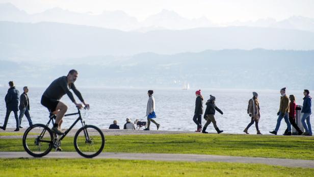Ein Mann fährt mit dem Fahrrad an einem See entlang, während andere Menschen spazieren gehen.