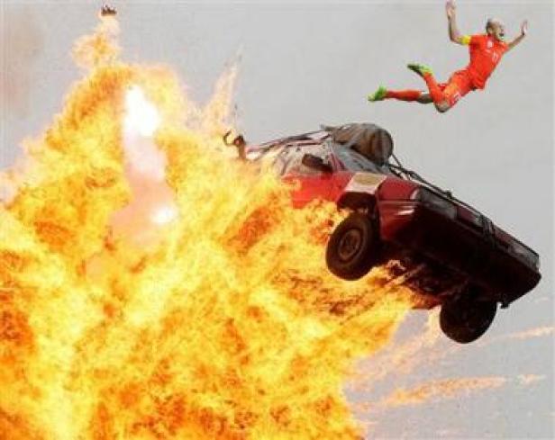 Ein Auto springt in die Luft, umgeben von einer großen Explosion, während eine Person in Sportkleidung daneben fliegt.