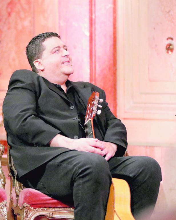 Ein Mann im schwarzen Anzug sitzt mit einer Gitarre auf einem Stuhl und schaut nach oben.