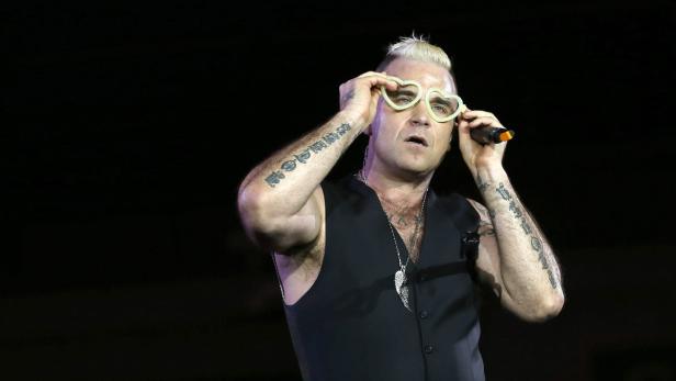 Robbie Williams mit herzförmiger Brille und Mikrofon auf der Bühne.