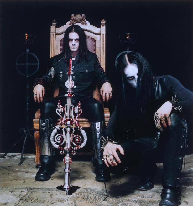 Zwei Männer mit Corpsepaint und dunkler Kleidung posieren vor einem dunklen Hintergrund.