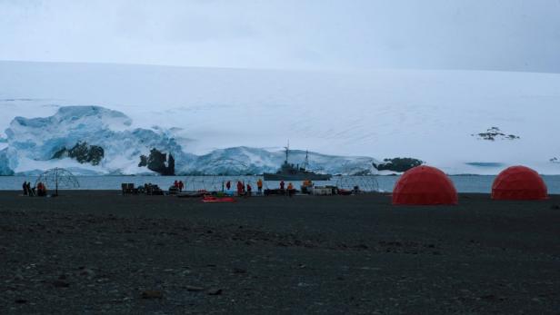 Eine Forschungsstation mit roten Kuppelzelten und einem Schiff in der Antarktis.