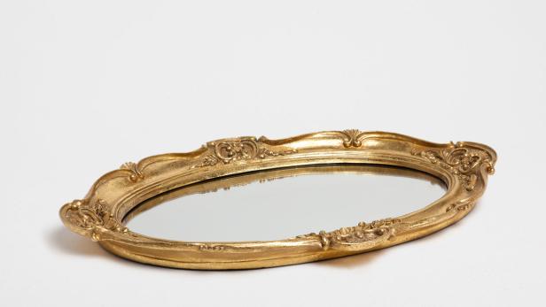 Ein ovaler Spiegel mit einem verzierten, goldenen Rahmen liegt auf einer weißen Oberfläche.