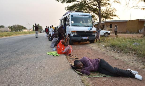 Eine Gruppe von Menschen wartet neben einem Bus am Straßenrand, einige sitzend oder liegend.